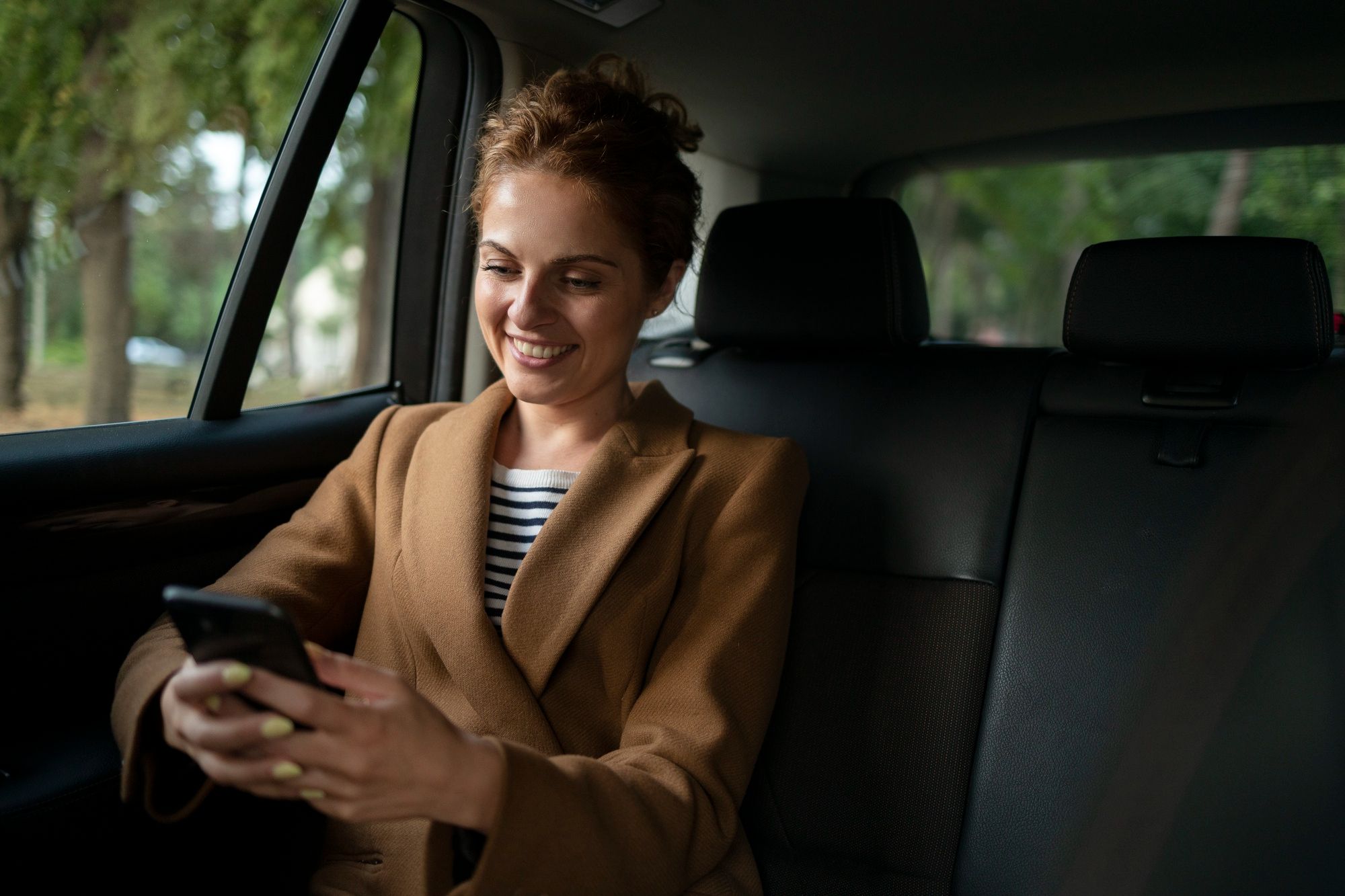 A woman smiles at her phone in the back seat of a car